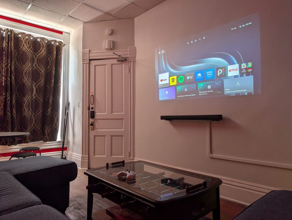 The projector in use in the Muskie Meeting room, showing streaming apps on the wall above a soundbar, with the foosball coffee table and an Xbox controller in the foreground.