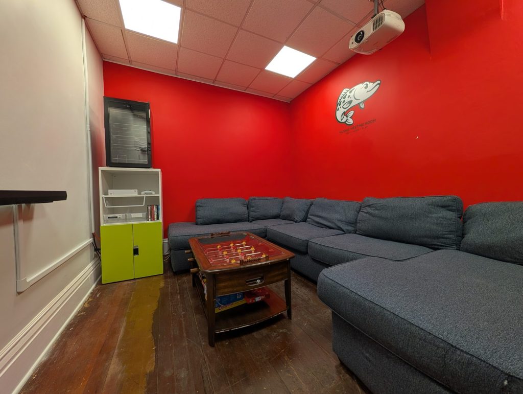 The Muskie Meeting room: a large grey sectional facing a foosball coffee table, with the Muskie Meeting Room logo on the red wall, a shelf of board games and an Xbox, and a ceiling-mounted projector.