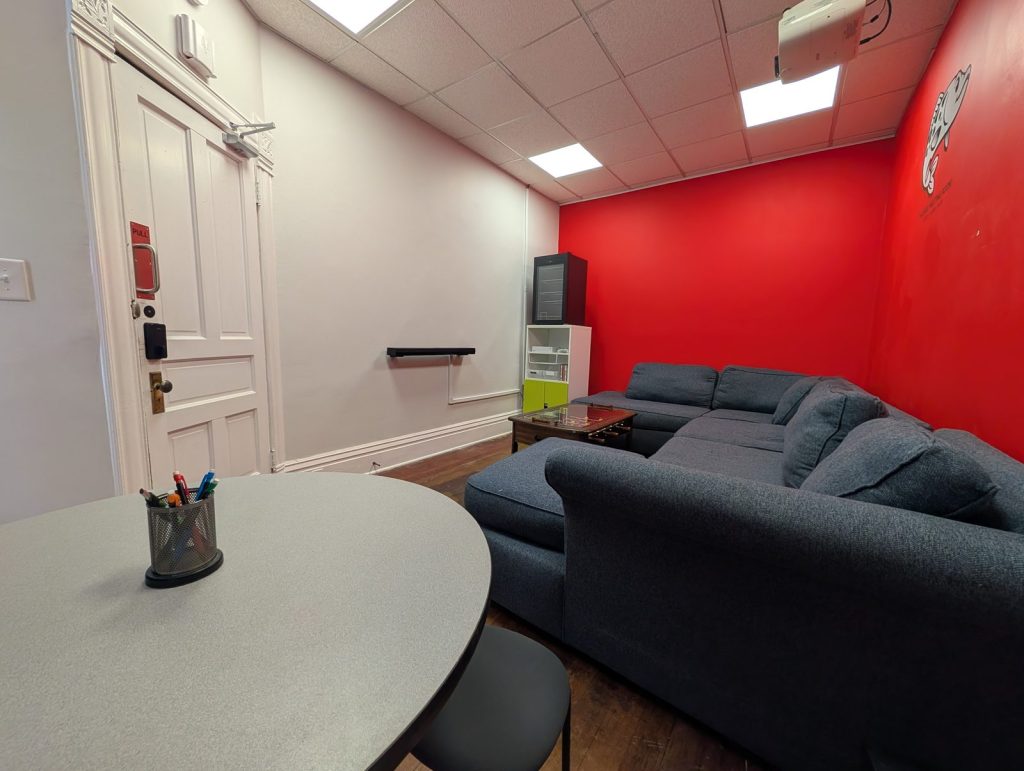 The Muskie Meeting room seen from the round work table, with dry-erase markers on the table, a whiteboard on the side wall, the grey sectional along the red logo wall, and the private entrance door.
